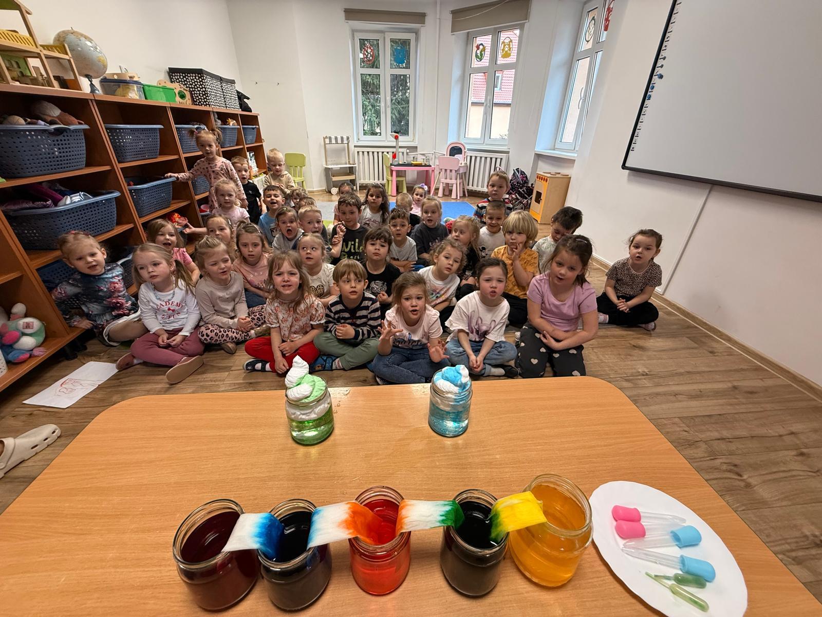 Dzieci siedzące na dywanie obserwują doświadczenia z wodą